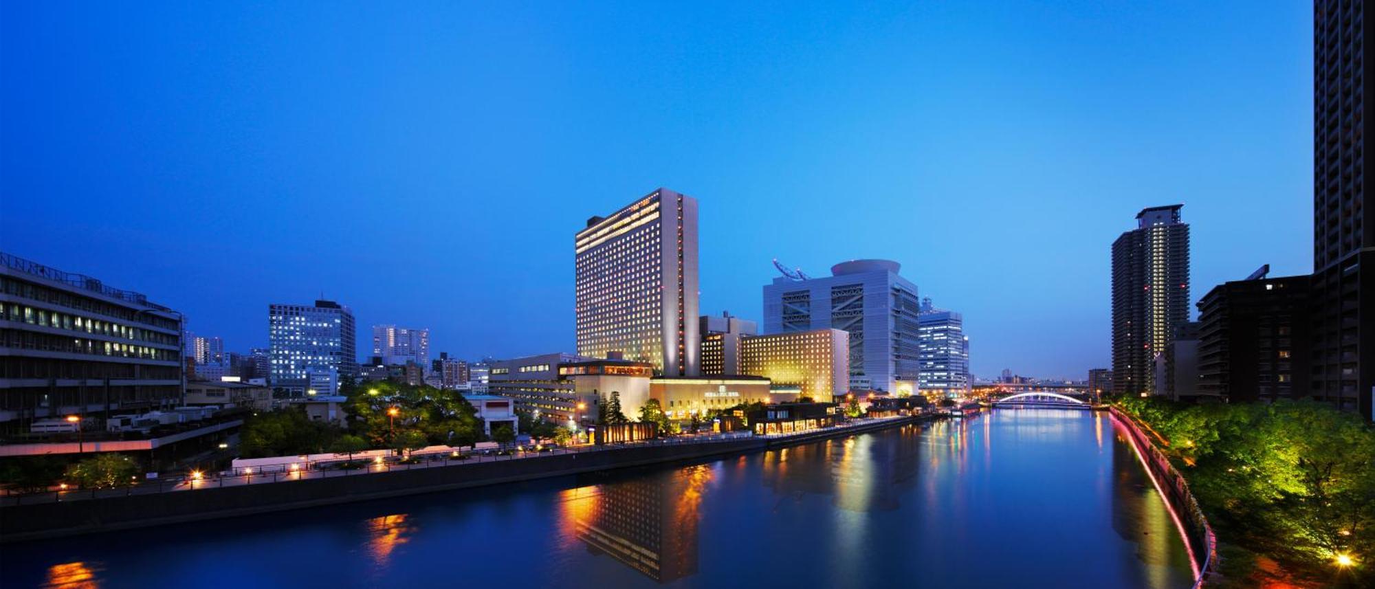 Rihga Royal Hotel Osaka, Vignette Collection By Ihg Exterior foto The photo shows a cityscape at dusk, featuring a modern hotel and several tall buildings along a river. The structures are illuminated with lights, reflecting beautifully on the water. The atmosphere has a serene and urban feel, with a clear blue sky